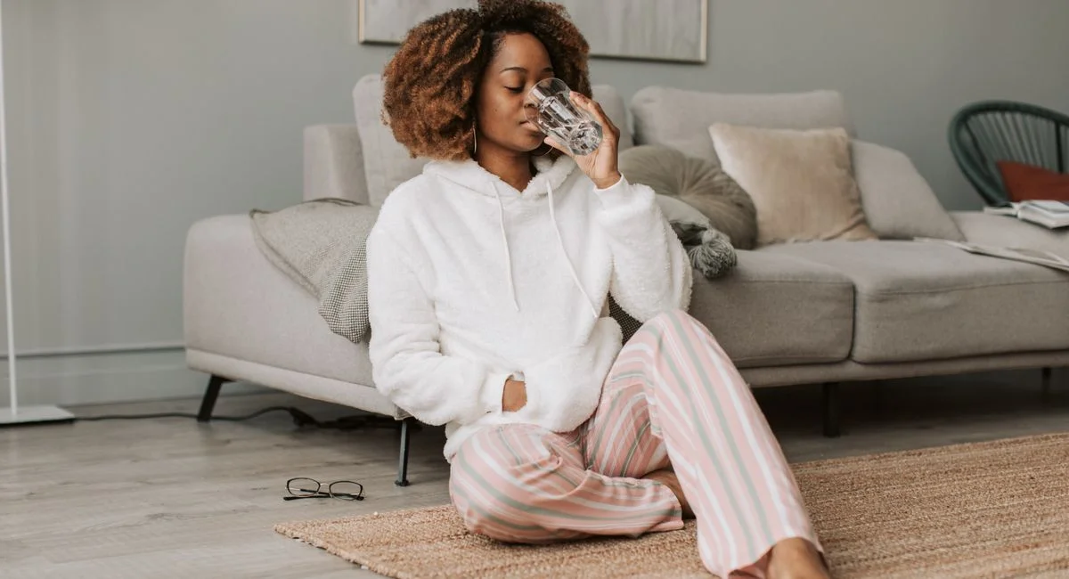 A woman in comfortable attire drinking a glass of water while sitting on a rug, embracing a morning routine.