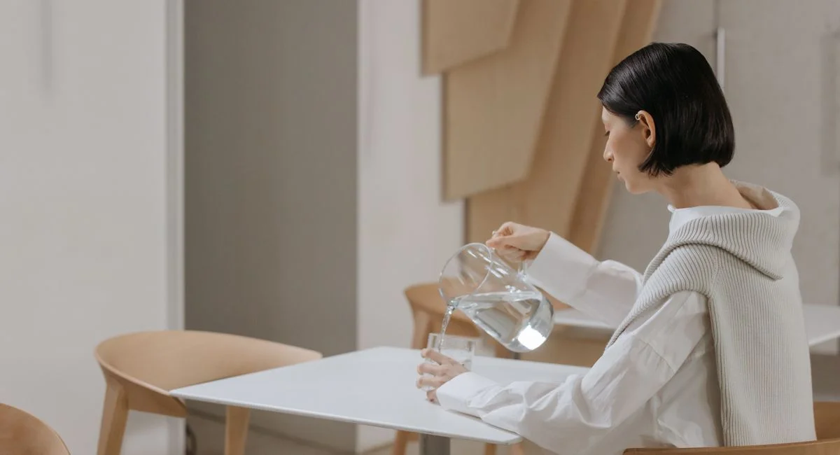 Adult woman in white pouring water into a glass on a minimalist table indoors.