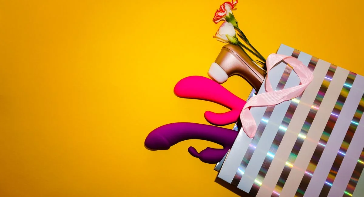 Colorful vibrators with flowers in a holographic gift bag, set against a bold yellow background.