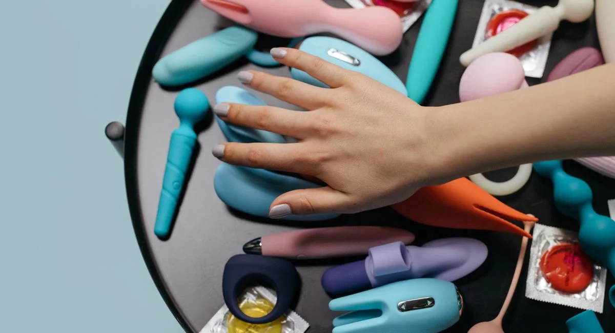 A hand reaching over a variety of colorful sex toys arranged on a table.
