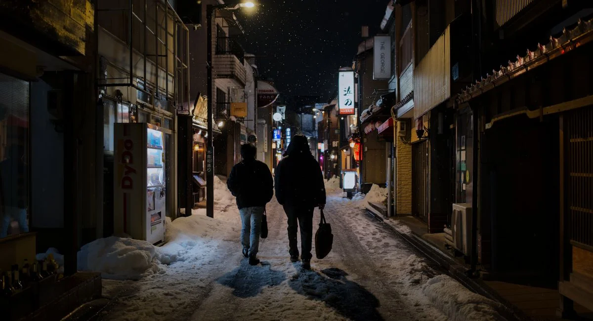 Two people walk through a snowy street in Kanazawa, Japan, surrounded by glowing lights and falling snow at night.