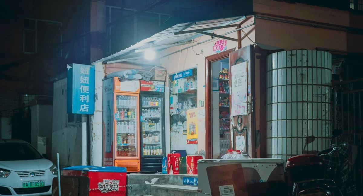 A small illuminated corner store on an urban street at night with visible goods.