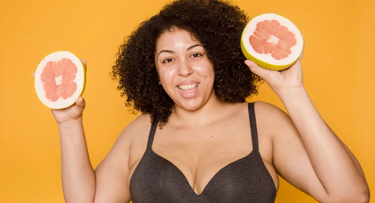 Cheerful plus size African American female in bra with curly hair and cut juicy grapefruit looking at camera on yellow backgr