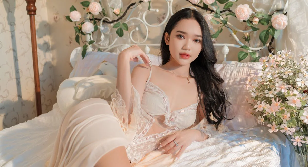 A woman in elegant lingerie poses gracefully on a floral-themed bed, surrounded by soft roses.