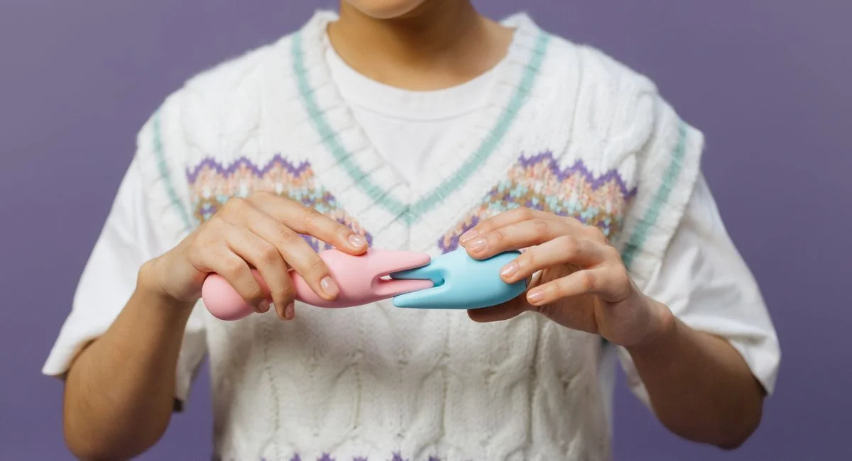 Hands holding pink and blue vibrators against a purple background, highlighting modern intimacy tools.