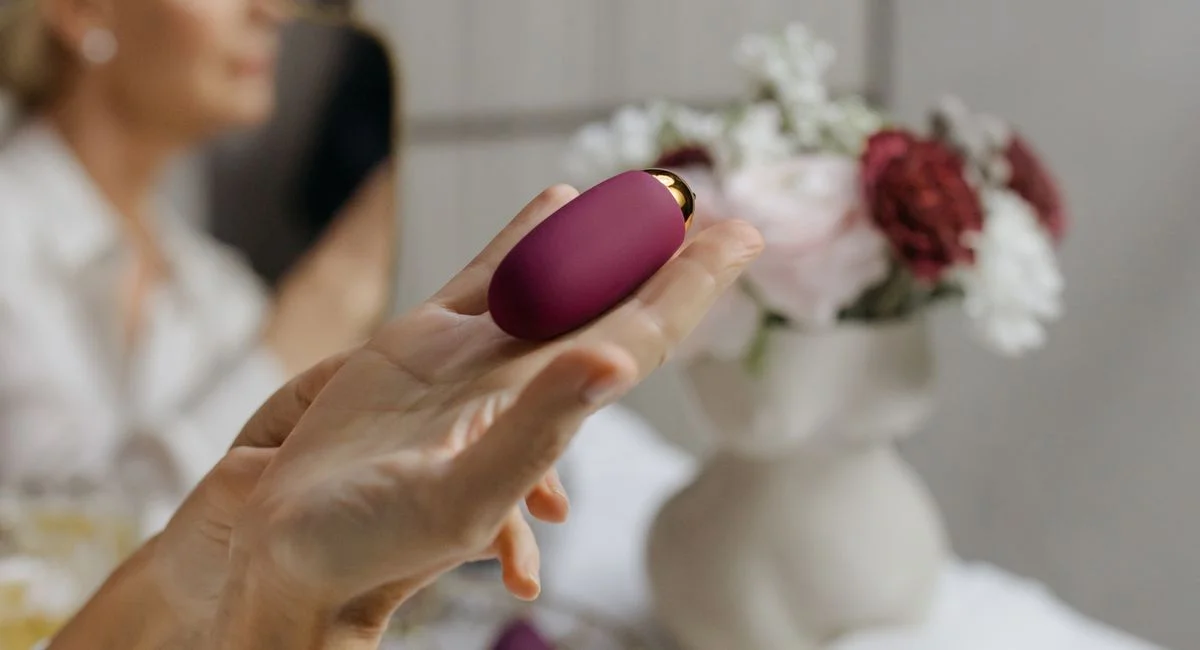 A mature woman elegantly holds a purple object near a floral arrangement in a reflective setting.